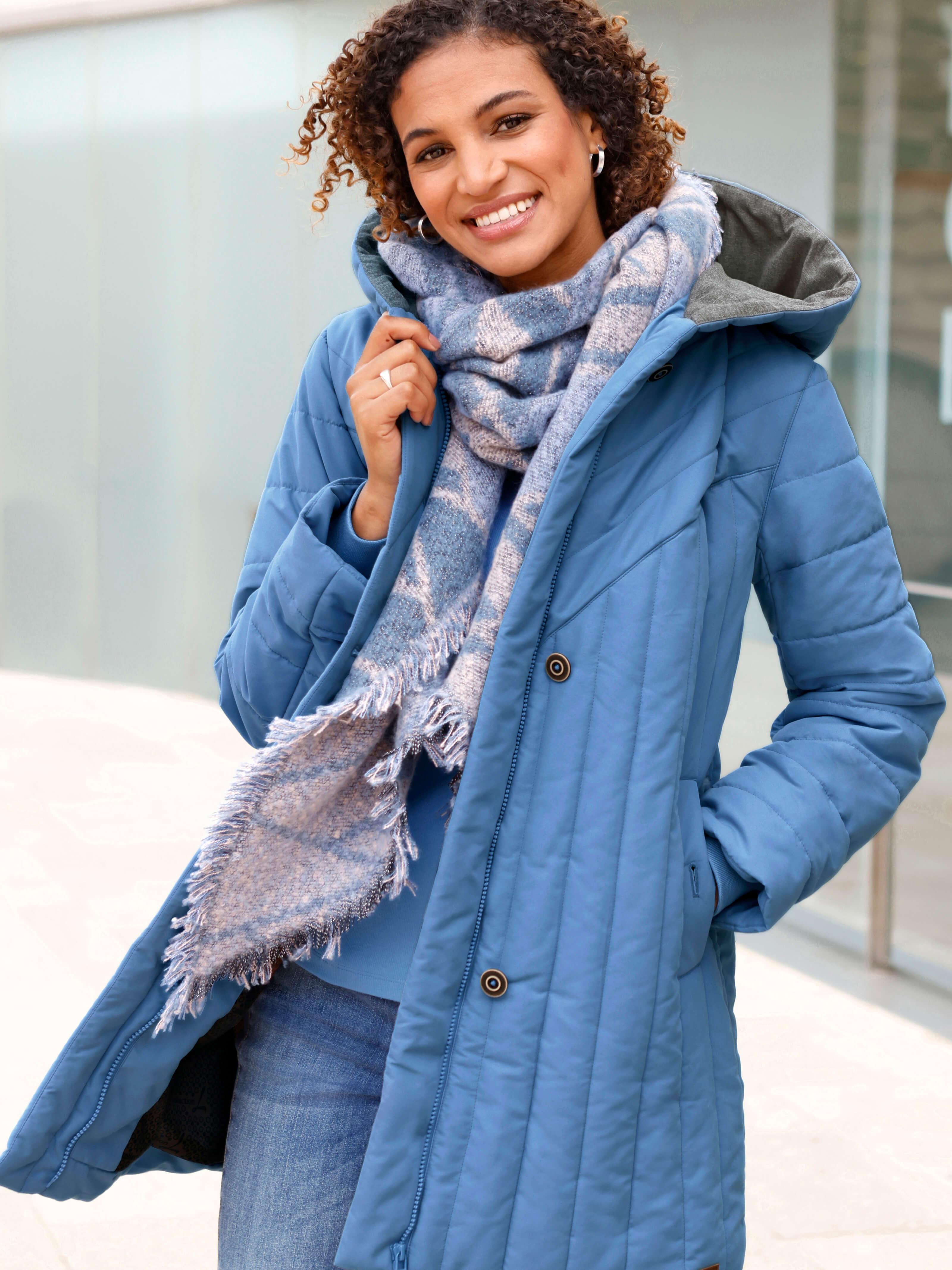Ein Model trägt einen blauen Steppmantel, Jeans und einen gemusterten Schal vor einem Gebäude.