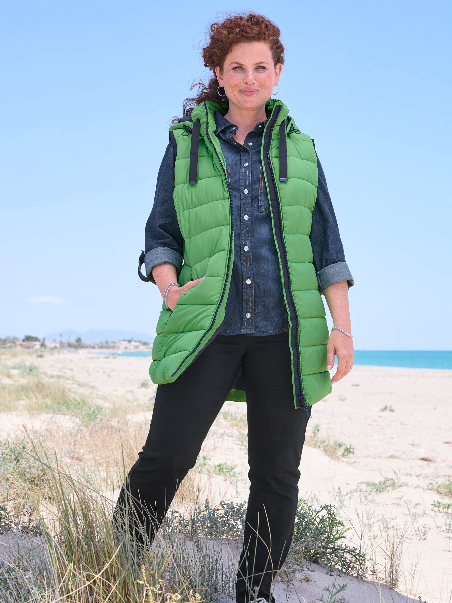 Eine Frau mit lockigem rotem Haar steht lächelnd am Strand. Sie trägt eine leuchtend grüne Kapuzenweste über einem Jeanshemd und einer schwarzen Hose. Im Hintergrund sind das Meer und eine Küstenlinie unter blauem Himmel zu sehen.
