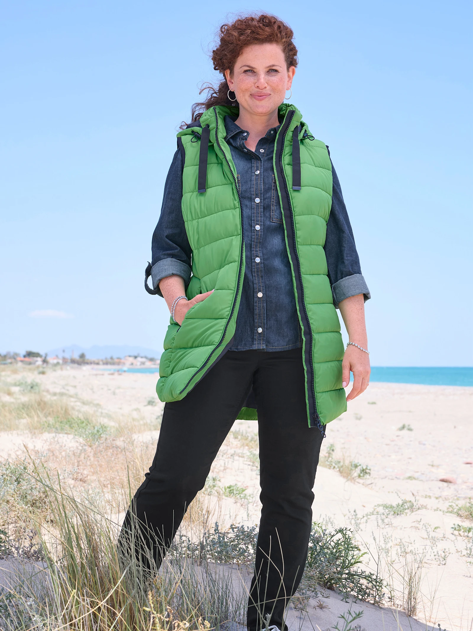 Eine Frau mit lockigem rotem Haar steht lächelnd am Strand. Sie trägt eine leuchtend grüne Kapuzenweste über einem Jeanshemd und einer schwarzen Hose. Im Hintergrund sind das Meer und eine Küstenlinie unter blauem Himmel zu sehen.