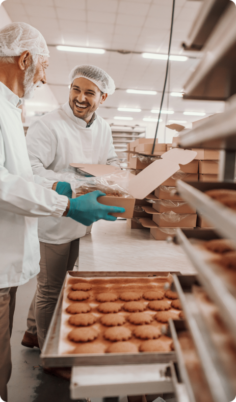 Two people inspecting biscuits on a conveyor belt in a food production facility, wearing protective clothing.