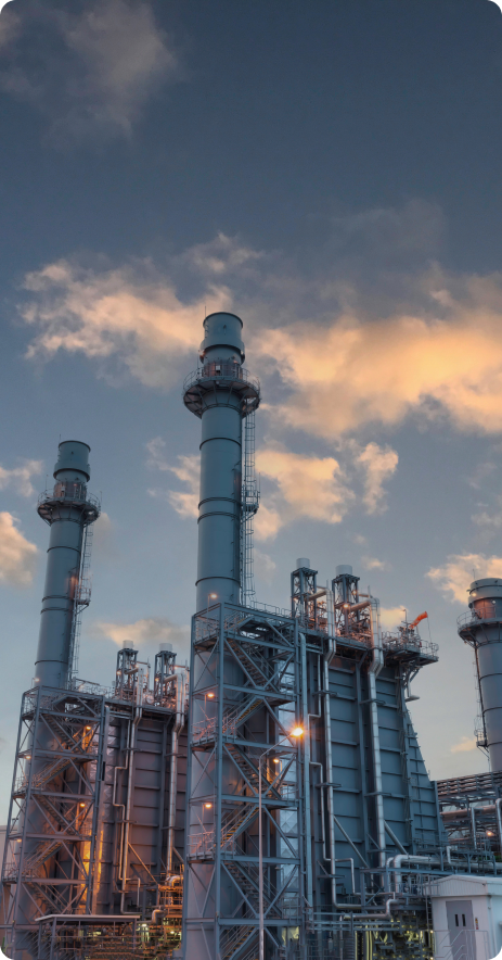 Industrial facility with tall metallic structures and chimneys. The sky is partly cloudy with hues of blue and orange.