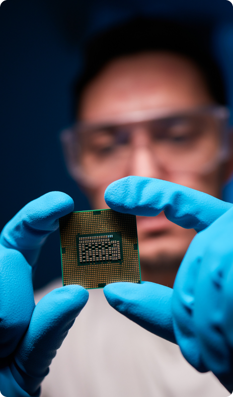 A close-up of gloved hands holding a square electronic chip, likely a CPU, against a blurred background.