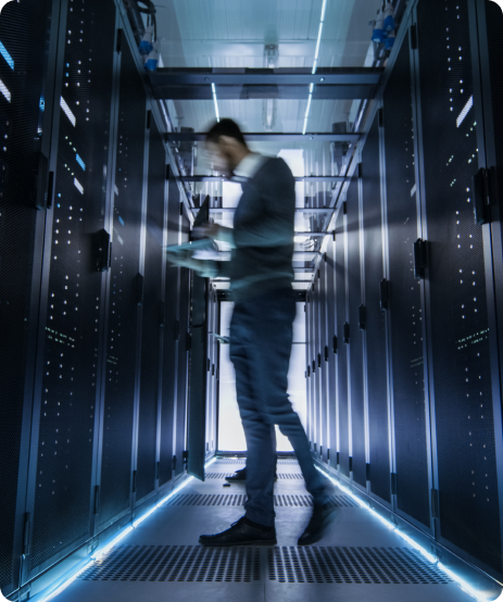 A person walks through a corridor of a data center filled with rows of server racks, holding a device. The environment is illuminated with blue lights.