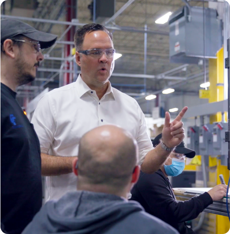 A man in a white shirt talks to three people in a factory setting. Two of them wear baseball caps, and one is sitting down. Machinery and industrial equipment are visible in the background.