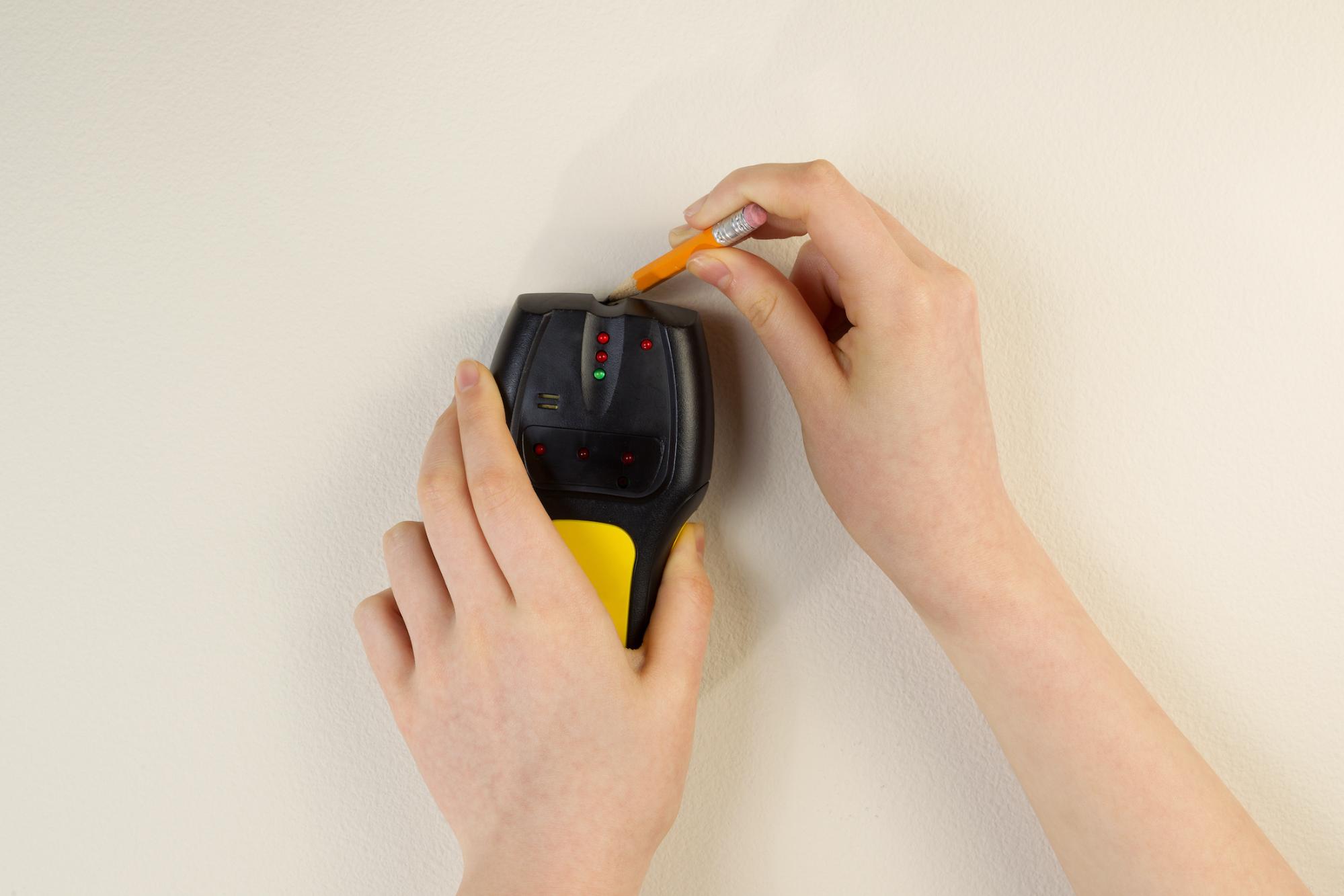 hands holding an electronic yellow and black stud finder using a pencil to mark location on a white wall 