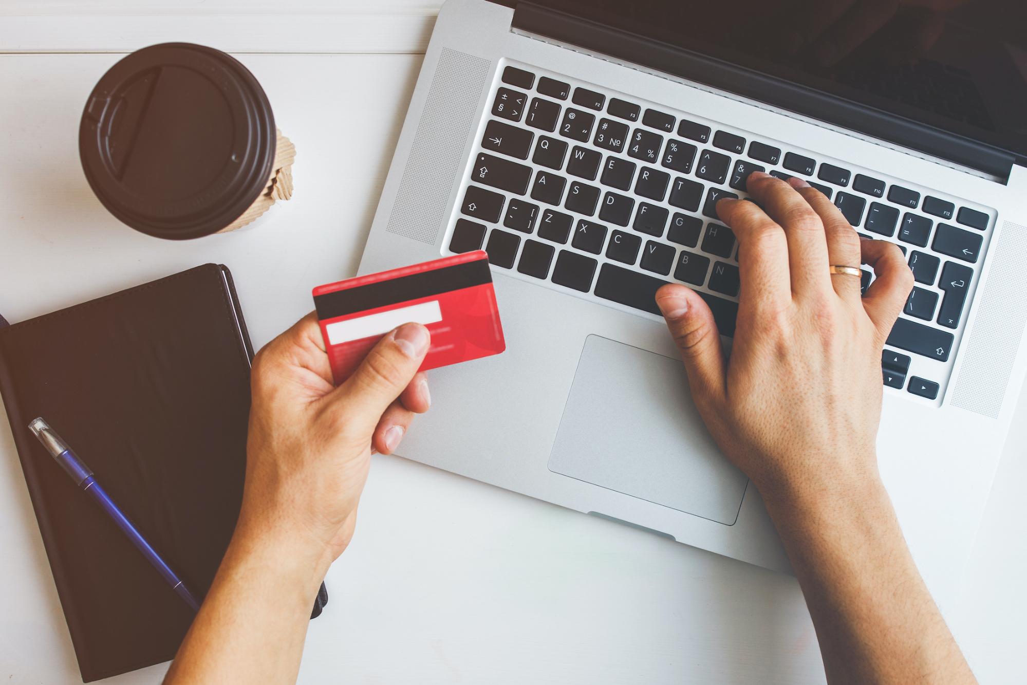 right hand of a person typing on an open laptop while the left hand is holding a red credit card