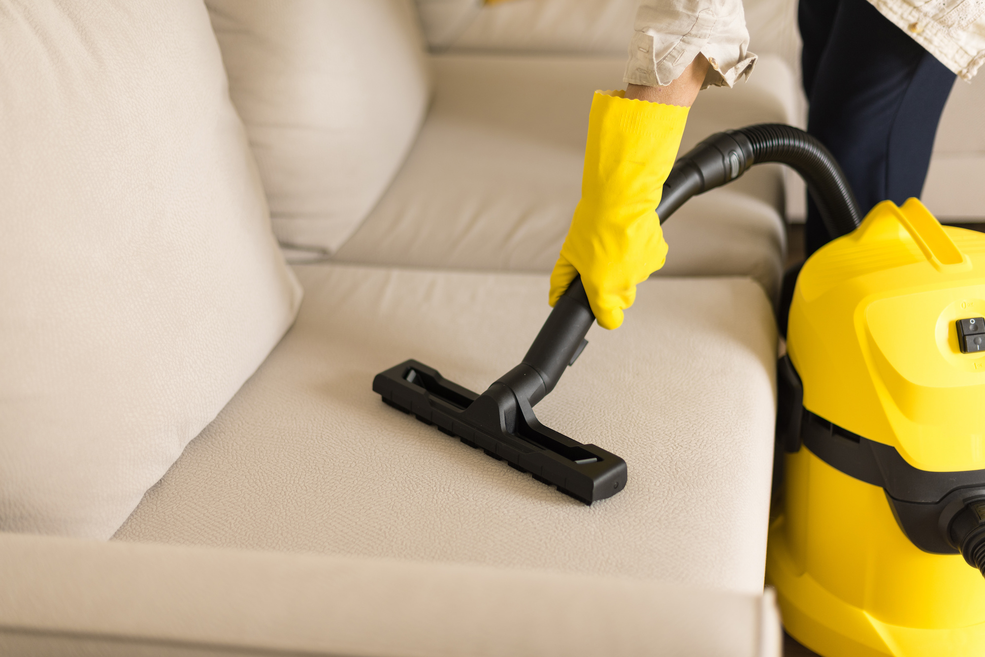 Hands in yellow cleaning gloves vacuum a grey couch.