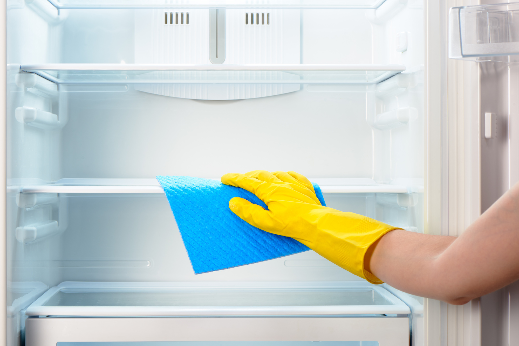 Hands with yellow gloves clean inner refrigerator shelves.