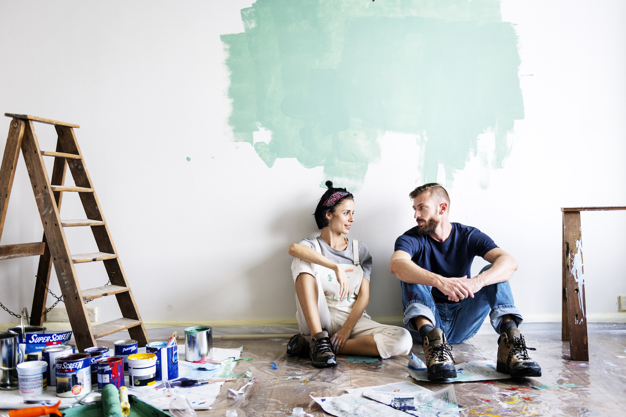 A man and woman sit in front of a partially painted wall in work clothes. 