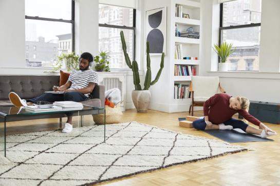 two individuals in a bright apartment. Black male is sitting on couch reading and white female is stretching on a yoga mat 