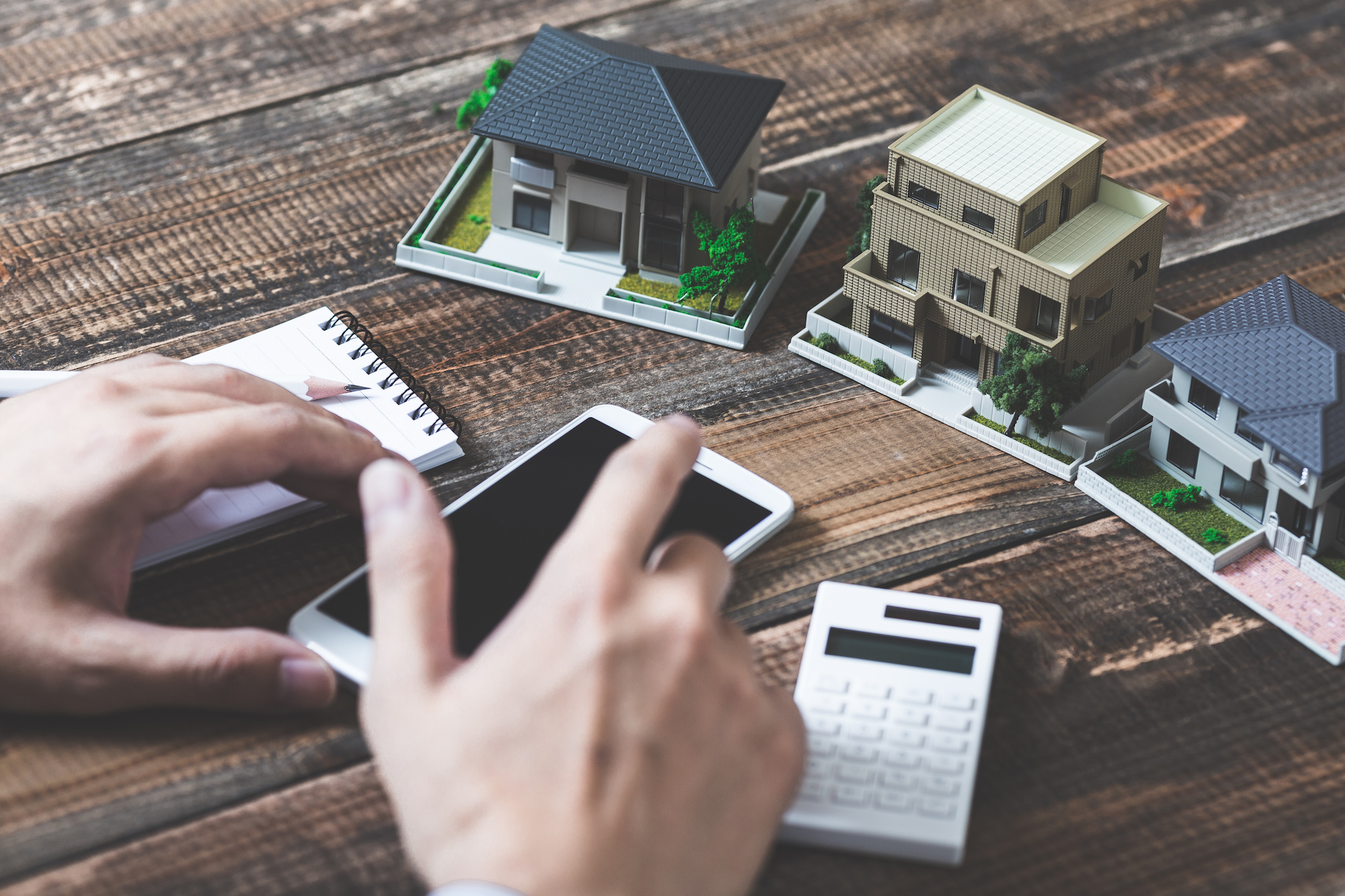 Three small miniature house models, a smartphone, calculator, notepad and person's hand with a pen.