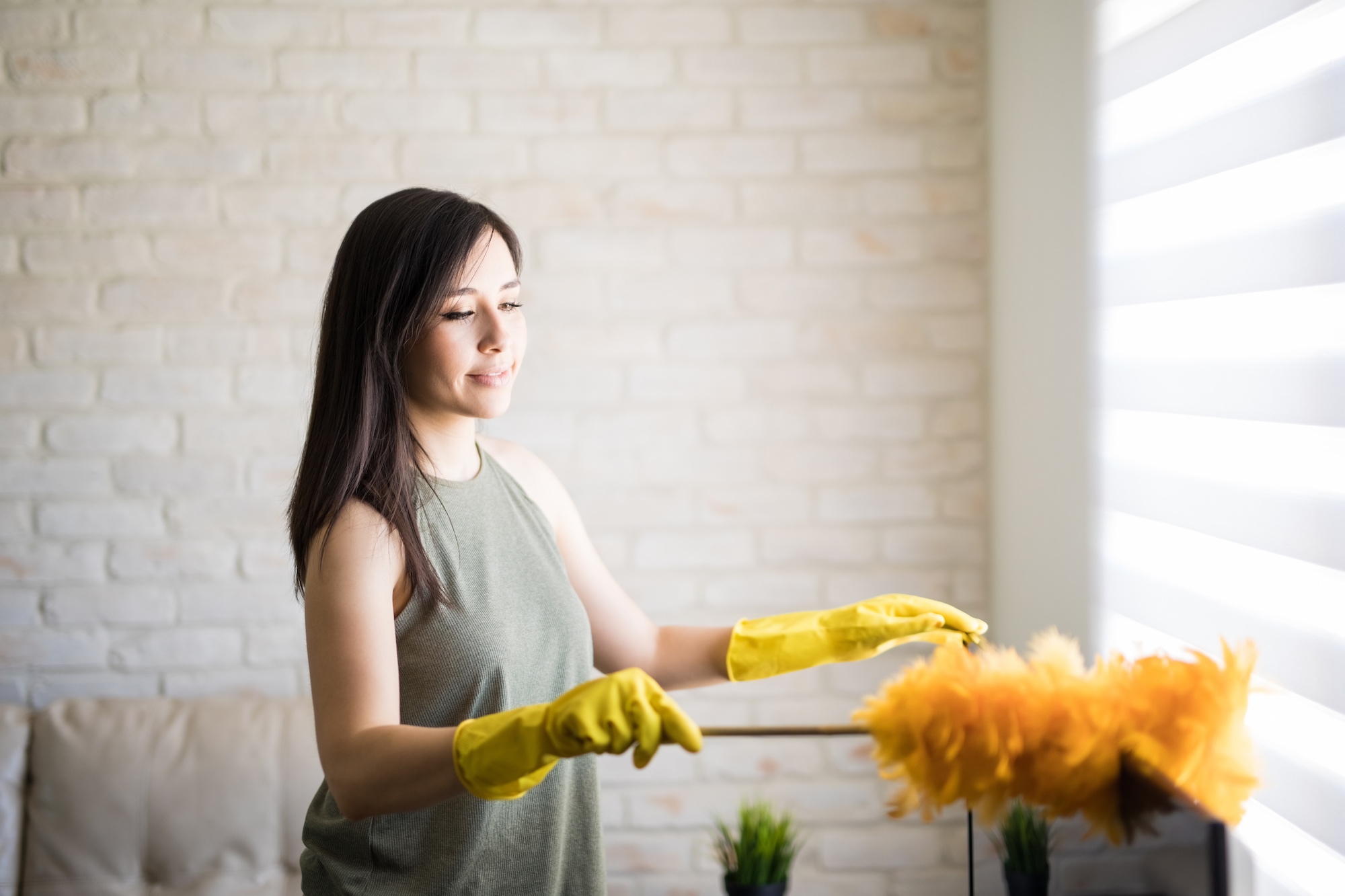Woman cleans blinds with a yellow duster and yellow cleaning gloves. 