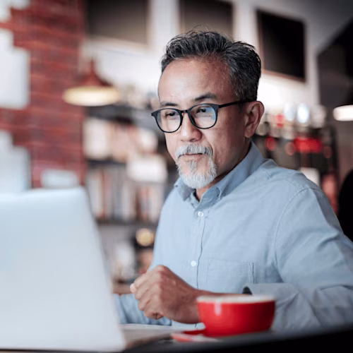 A man in a blue shirt uses a laptop at a café, with a red cup beside him, focusing on Docusign tasks.