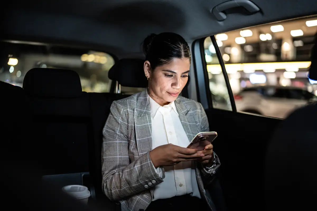 Una persona con un blazer a cuadros está sentada en un auto, concentrada en su teléfono inteligente, con luces borrosas de fondo.