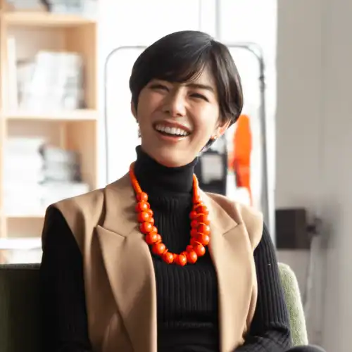 A professional Asian woman wearing an orange necklace and a tan vest is smiling in an office