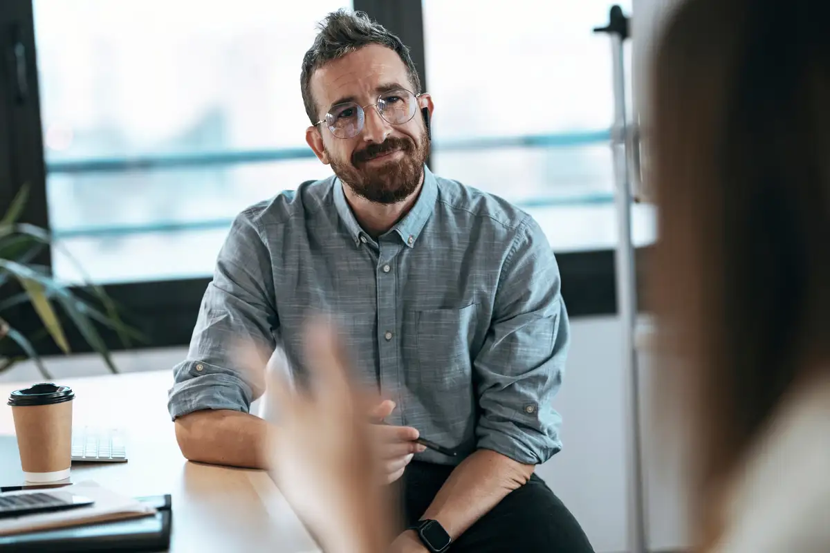 Een man in een blauw shirt zit aan een tafel en luistert aandachtig naar een vrouw die gebaren maakt tijdens een gesprek.