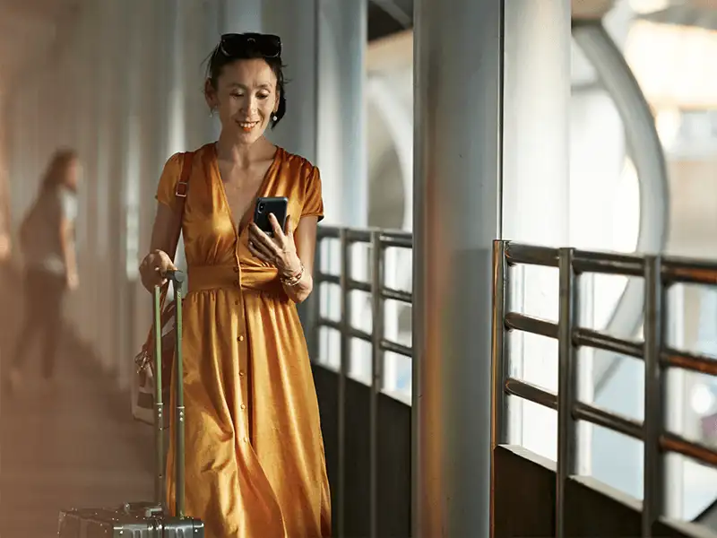 Woman in an orange dress walking with luggage on a covered bridge looking at her mobile device