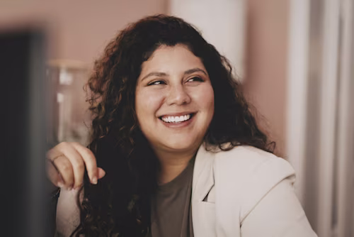 Una persona con cabello rizado que lleva un blazer claro está sentada en un escritorio, apoyando su barbilla en su mano, en una habitación suavemente iluminada.