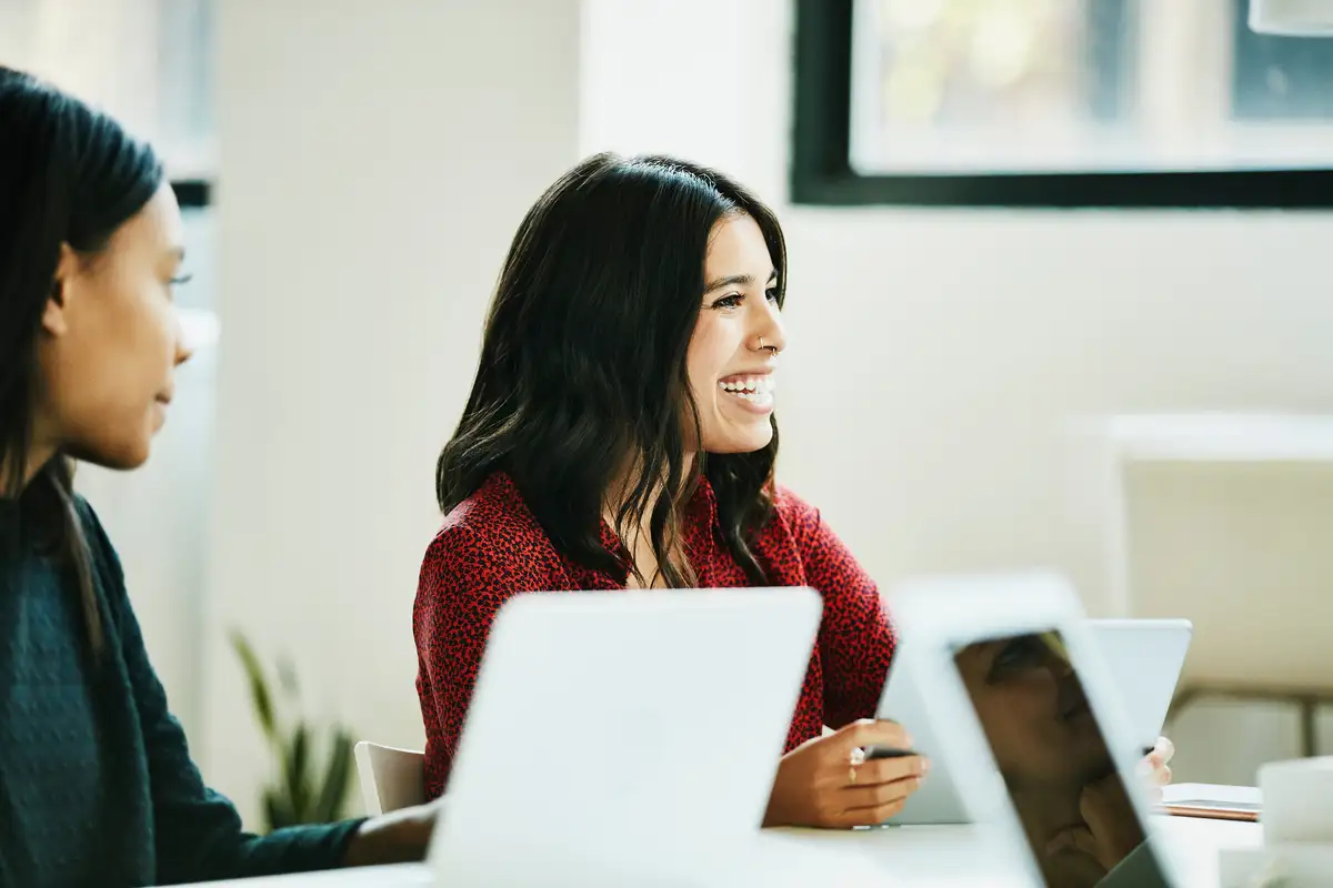Una mujer con una camisa roja de estampado se sienta en una mesa con laptops y una tableta, participando en una conversación.