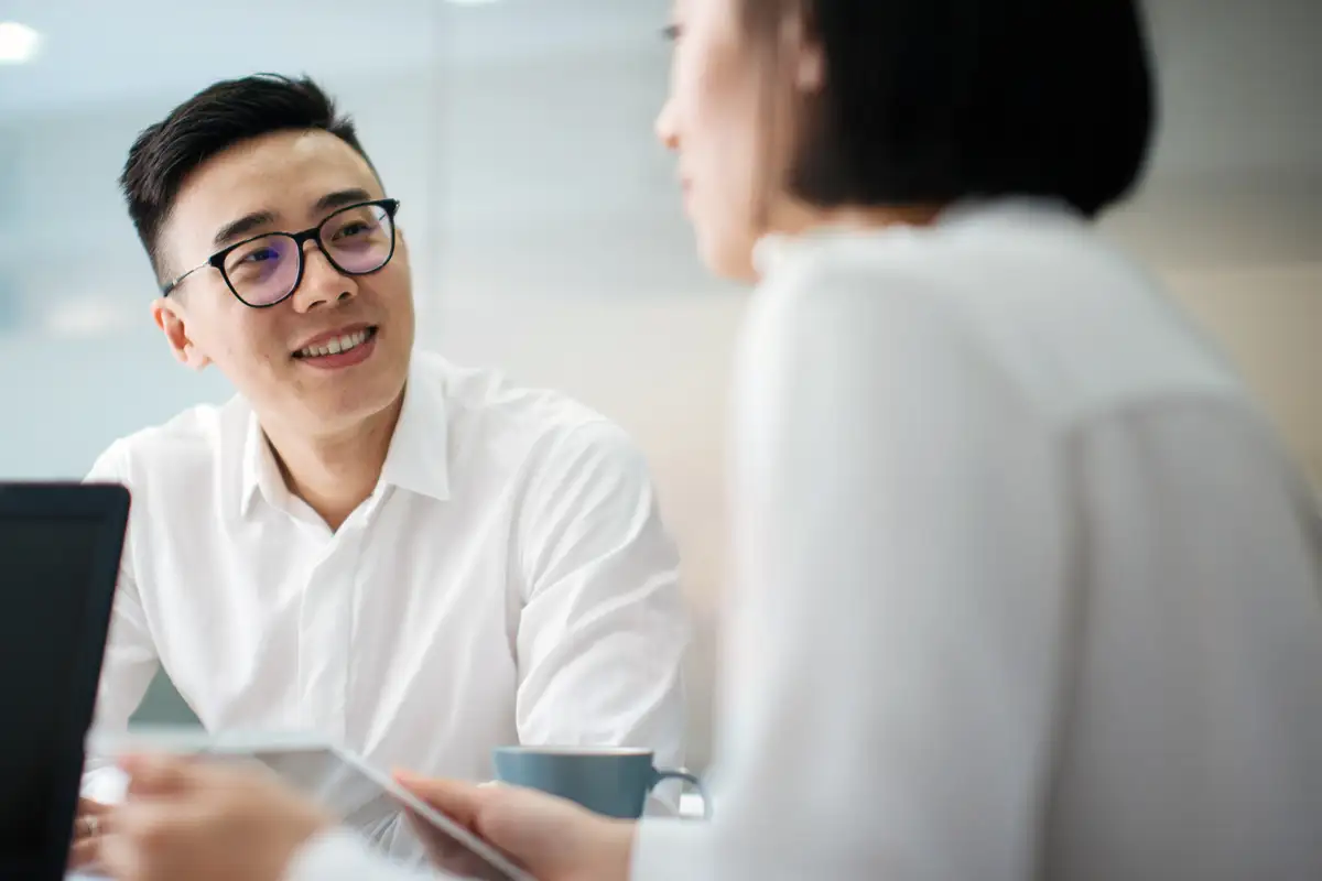 二人の女性が明るいオフィスのデスクでコンピュータとタブレットを使って協力している一方で、二人の男性が背景で作業しています。