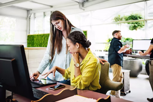 Twee vrouwen werken samen aan een bureau met een computer en tablet, terwijl twee mannen op de achtergrond in een lichte kantoorruimte werken.