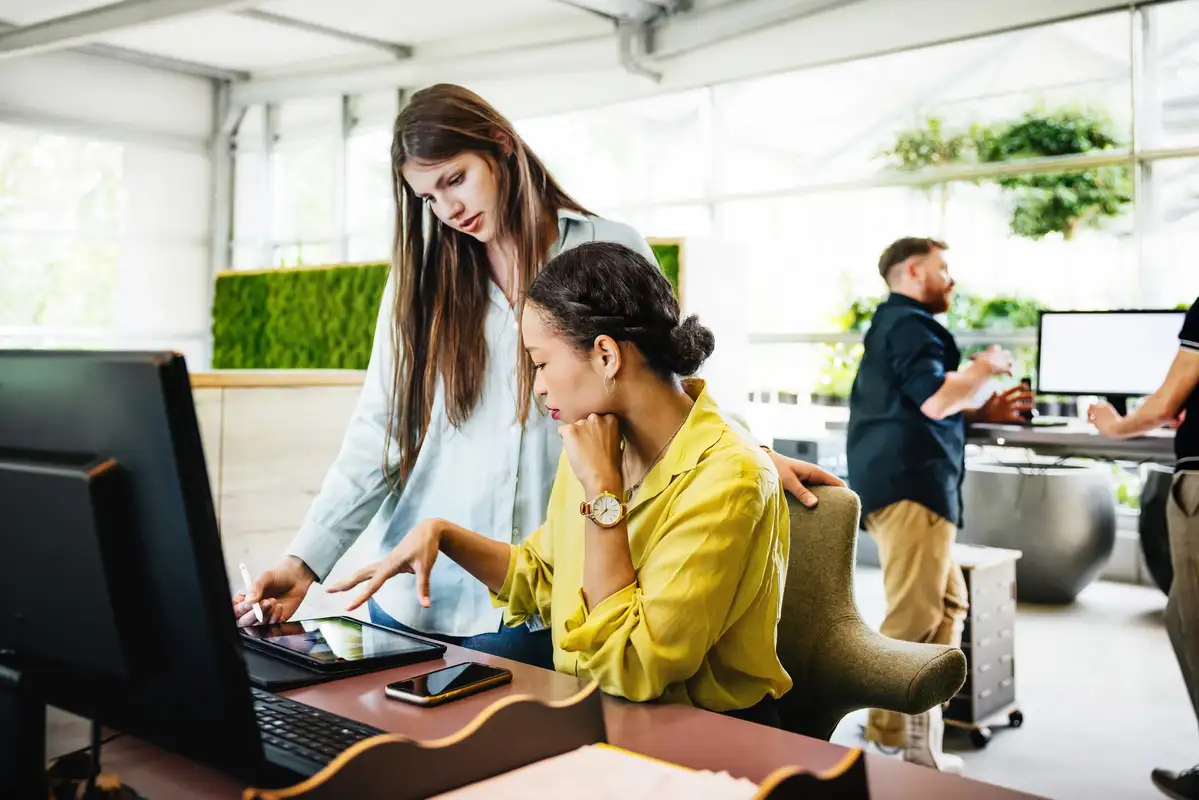 Twee vrouwen werken samen aan een bureau met een computer en tablet, terwijl twee mannen op de achtergrond in een lichte kantoorruimte werken.
