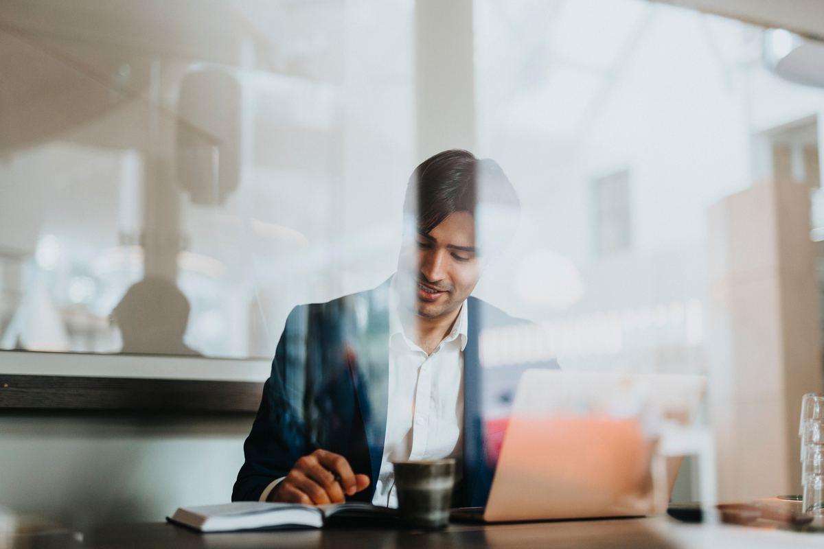 Un hombre con traje está sentado en una mesa con un portátil, una libreta y un café, trabajando en un ambiente de cafetería moderno y luminoso.