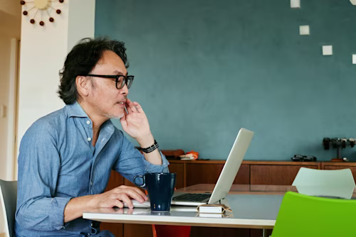 薄い青いシャツを着た男性が会議テーブルでタブレットを持っており、背景には女性とノートパソコンが見えます。