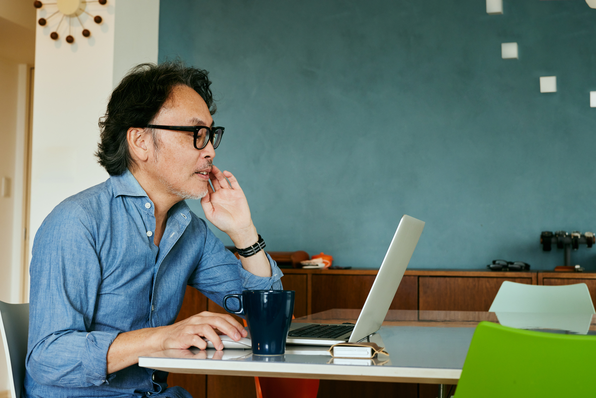 薄い青いシャツを着た男性が会議テーブルでタブレットを持っており、背景には女性とノートパソコンが見えます。