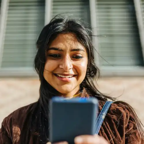 Een vrouw in een corduroy jasje glimlacht terwijl ze naar haar mobiele telefoon kijkt buiten een gebouw van bakstenen