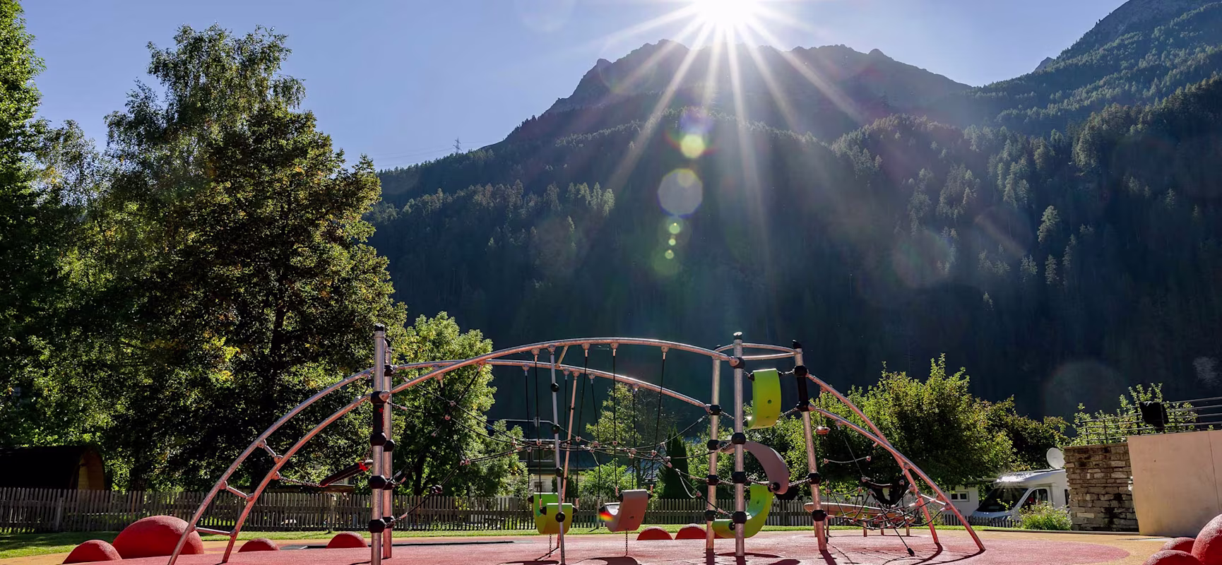Sunlit mountain playground with colourful climbing frame on red rubber ground, surrounded by trees and wooden fences.