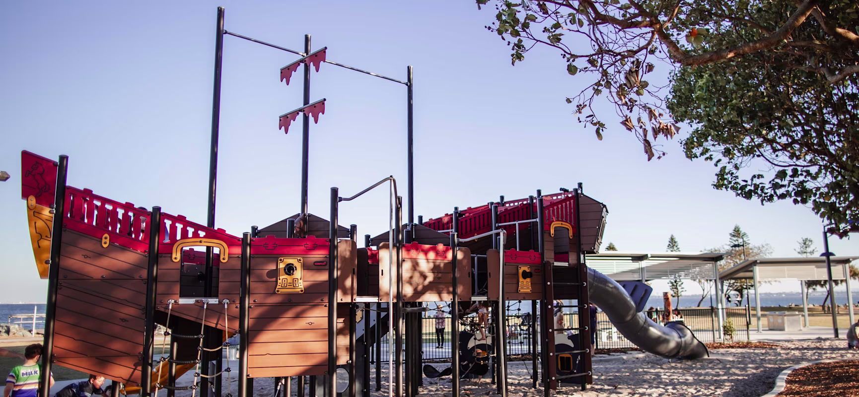 Children play on a large pirate ship playground in sandy park, with slides and climbing nets under clear blue sky and nearby trees.
