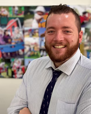 Person in a striped shirt and tie stands with folded arms in front of a colorful photo collage on a wall.