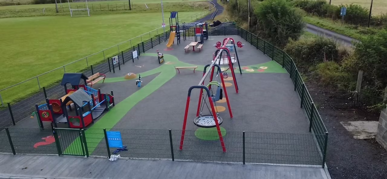 Fenced outdoor playground with swings, slides and colourful climbing structures beside a grass sports field and rural road