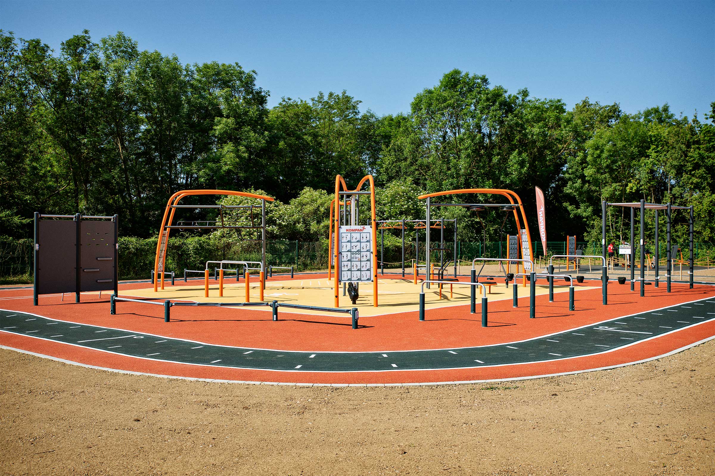 Outdoor fitness park with orange and black exercise structures and a running track, surrounded by trees under a clear blue sky.
