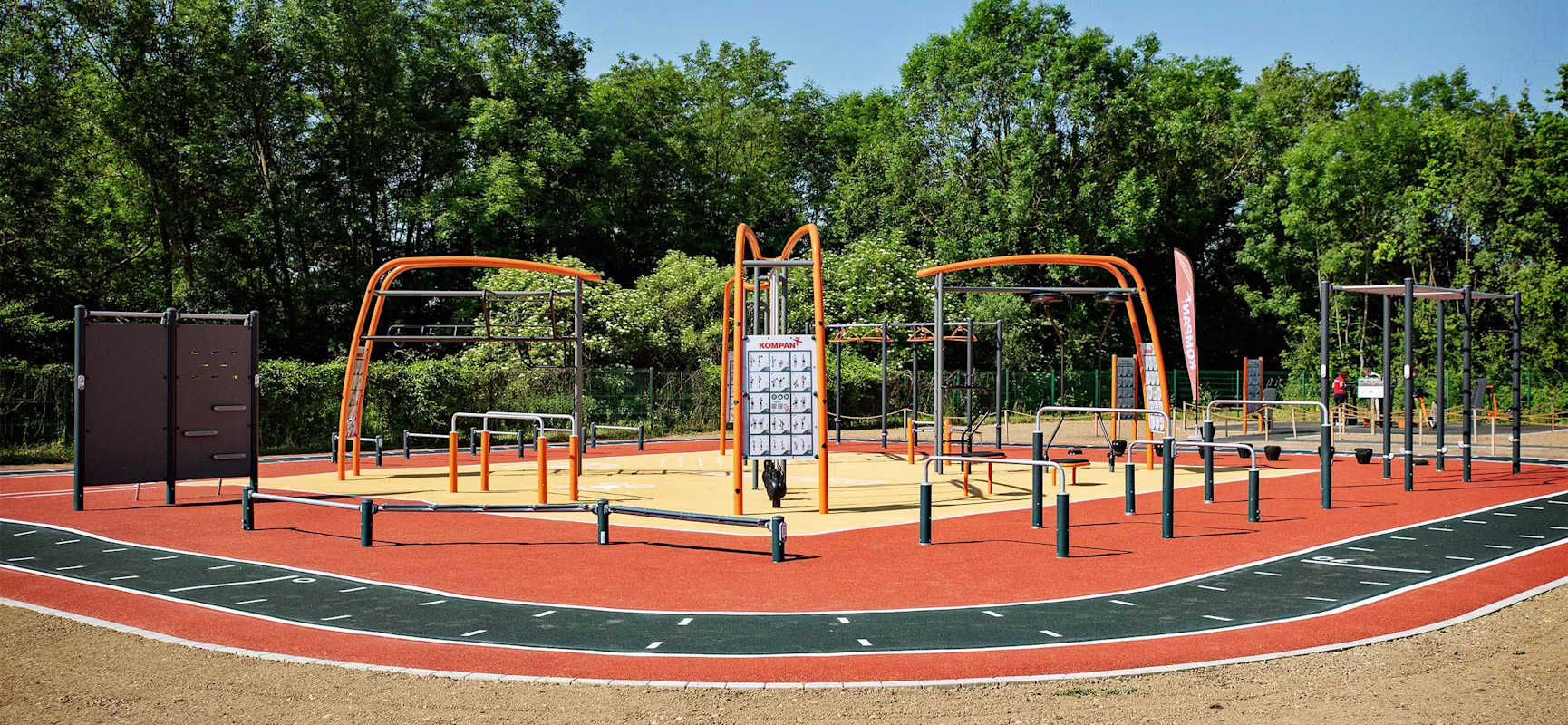 Outdoor fitness park with orange and black exercise structures and a running track, surrounded by trees under a clear blue sky.