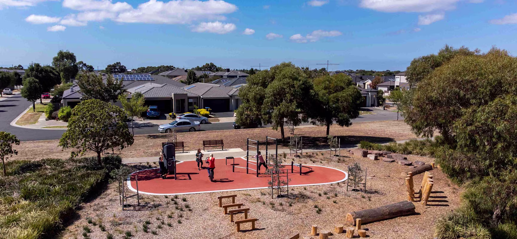 Aerial view of a small suburban playground with people on red rubber flooring, surrounded by trees, logs and nearby houses under blue sky