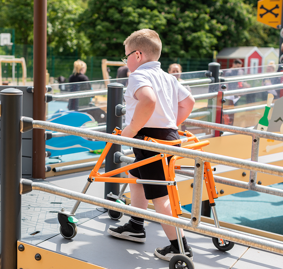 Child using a yellow walker on an accessible playground, wearing leg braces and a cap, with trees and equipment in the background.