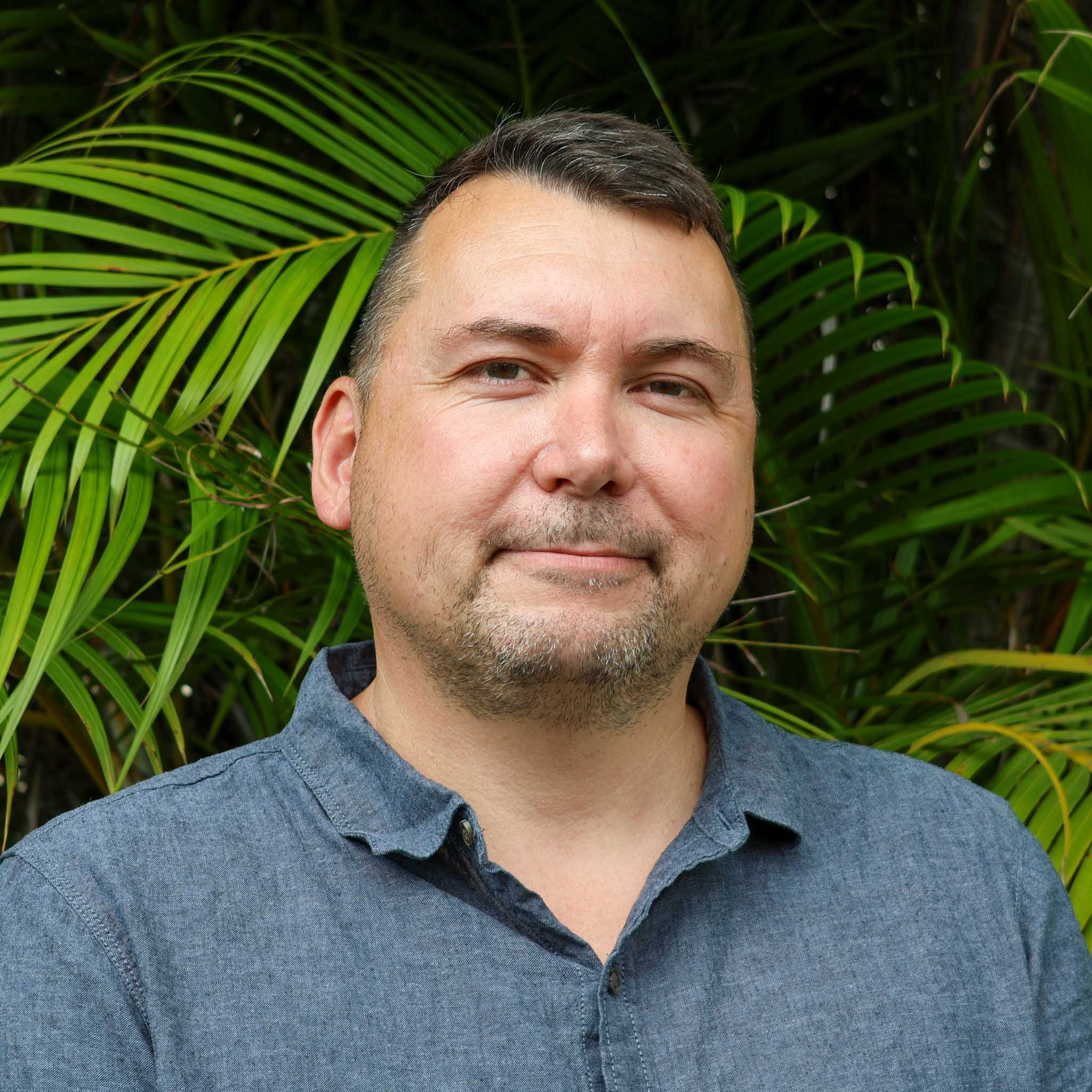 Person with blurred face in a blue button-up shirt standing in front of lush green palm leaves outdoors