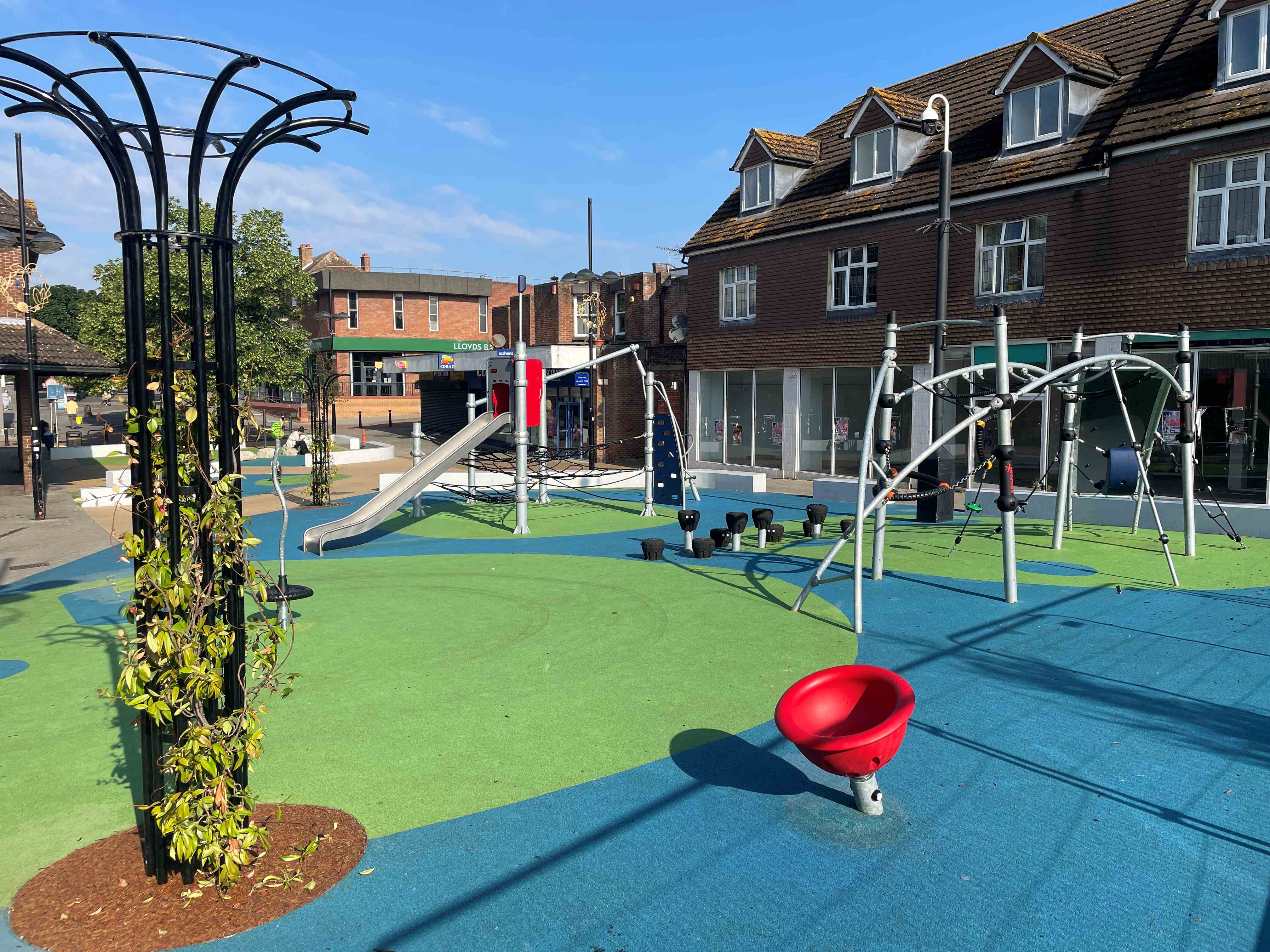 Sunny town playground with colourful rubber flooring, slides and climbing frames, surrounded by brick shops and a few trees.