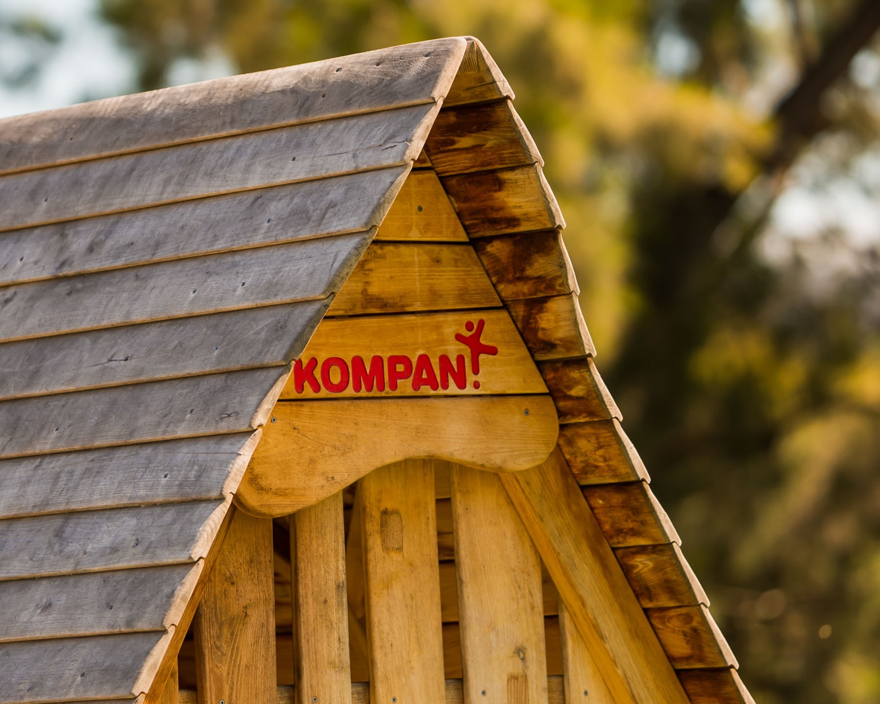 Close-up of a wooden playhouse roof with the brand name “KOMPAN” in red, set against a blurred green outdoor background.
