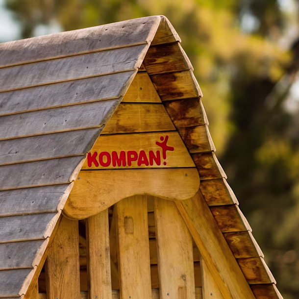 Close-up of a wooden playhouse roof with the brand name “KOMPAN” in red, set against a blurred green outdoor background.