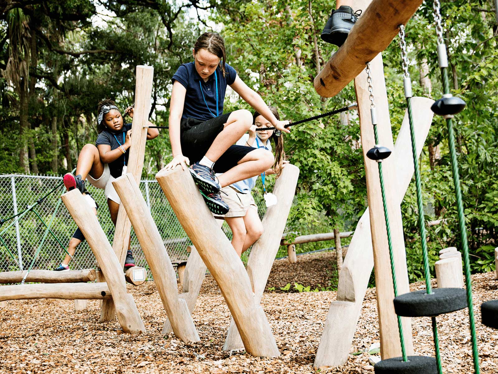 Children climbing wooden balance structures on an outdoor playground surrounded by trees and wood chips