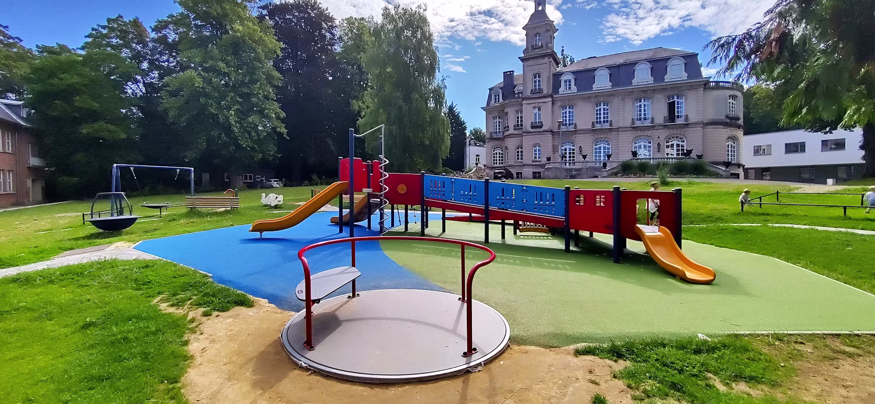 Colourful playground with slides, roundabout and swings on green lawn, set before a large historic building under a bright partly cloudy sky