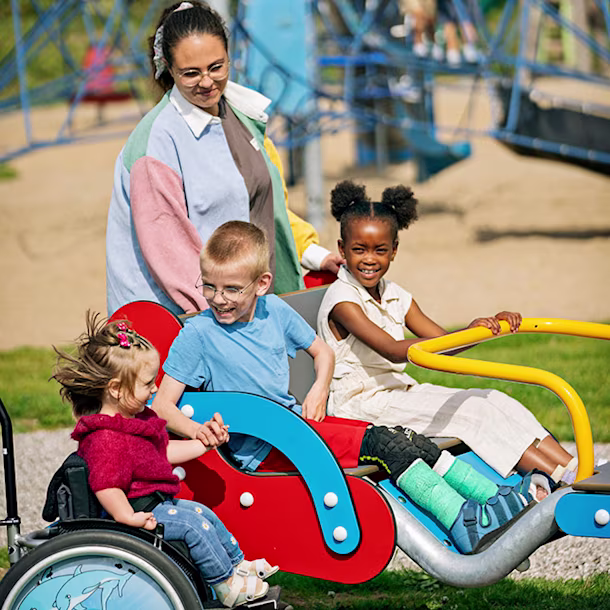 Children with and without wheelchairs ride an inclusive playground seesaw, pushed by an adult, with climbing frames in the background