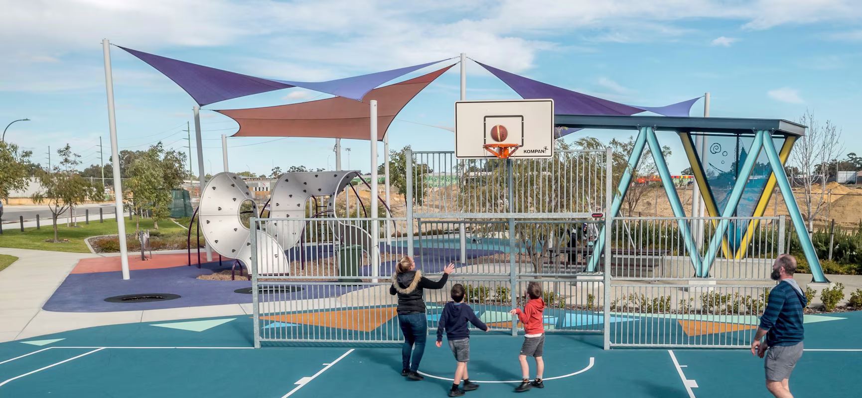 Family plays basketball on a blue outdoor court beside a colourful shaded playground with modern climbing structures.
