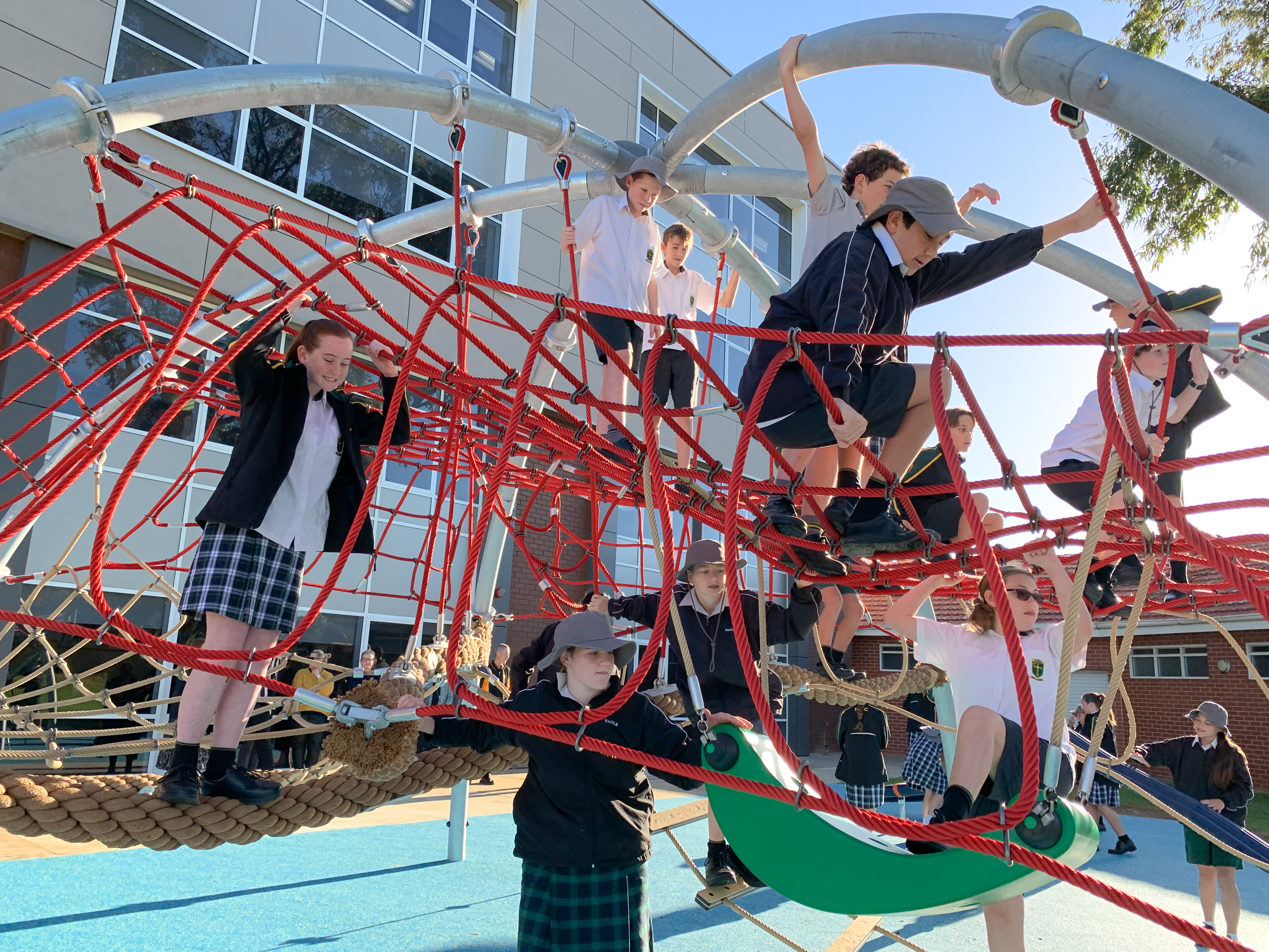Schoolchildren climbing a red rope playground structure outdoors, with a modern school building in the background.