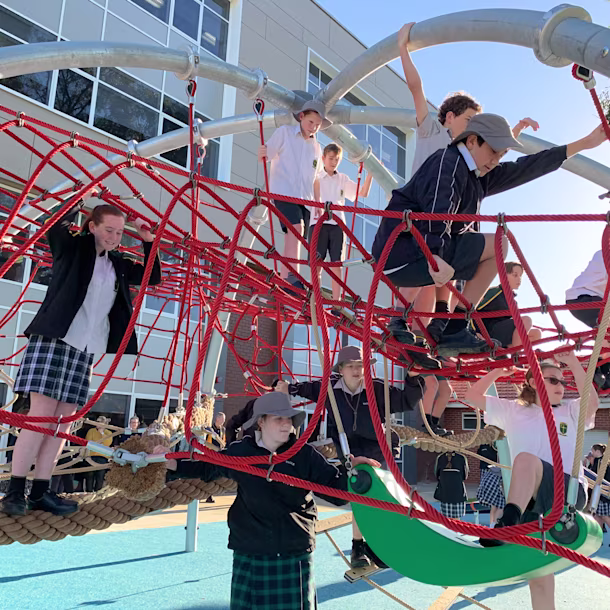 Schoolchildren climbing a red rope playground structure outdoors, with a modern school building in the background.