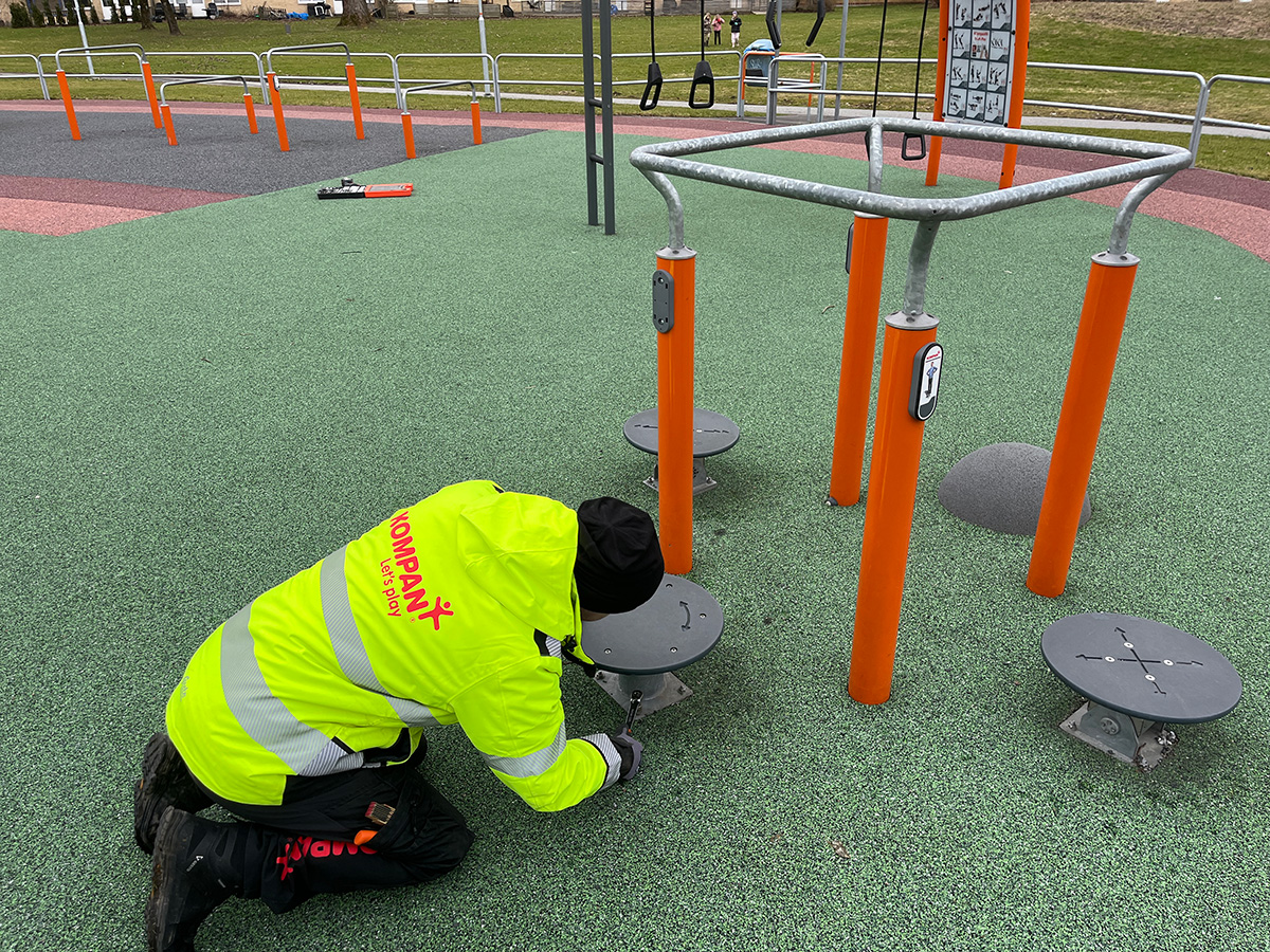 Worker in neon jacket kneels on a rubber playground, installing grey stepping platforms beside orange metal exercise bars.