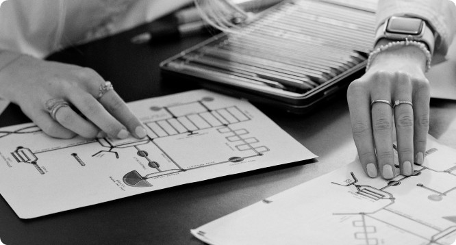 Close-up of two hands reviewing technical diagrams on paper, with a tray of drawing tools in the background.